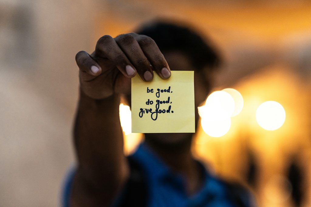A person holds a sticky note with an encouraging message to do good and thereby glorify God.