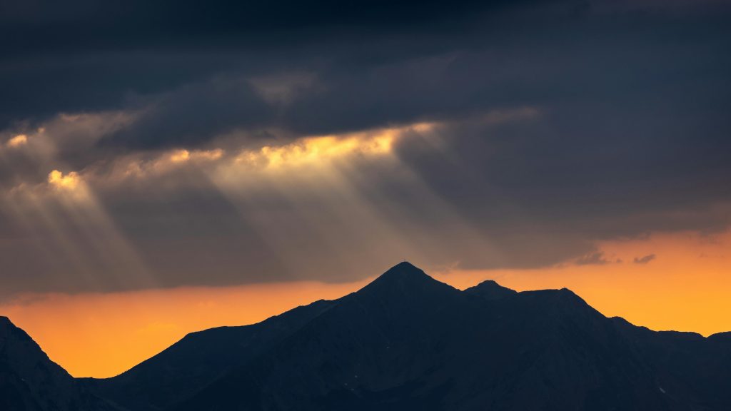 Sun rays pierce through clouds over mountains at sunset, creating a dramatic landscape scene.